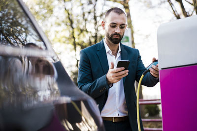 Businessman looking at smart phone holding electric car charging plug