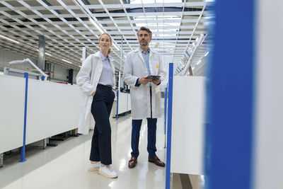 Engineer with hands in pockets standing by colleague holding tablet pc at factory