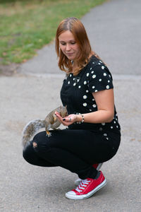 Beautiful young woman feeding squirrel while crouching on road