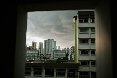 View of buildings against cloudy sky in city seen through window