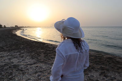 Rear view of woman wearing hat standing at beach during sunset