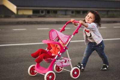 Cute girl with doll in baby stroller walking on road