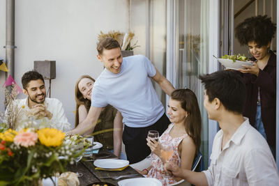 Smiling man and woman serving food to friends sitting at dining table during party in balcony
