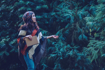 Side view of woman with book standing by trees in forest