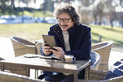 Happy man on video call using tablet pc sitting at sidewalk cafe