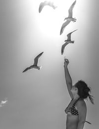 Side view of woman feeding seagulls while wearing bikini top against sky during summer