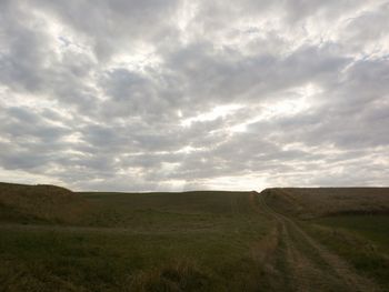 Dirt road passing through field against cloudy sky