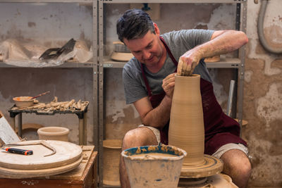 Full body of concentrated male master in apron sitting at table while sculpting with brown clay on throwing wheel