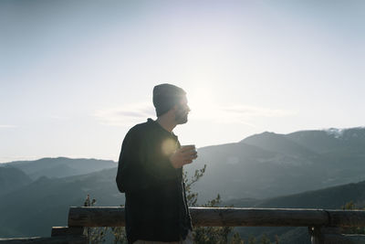 Side view of man looking at mountains against sky having a cup of coffee