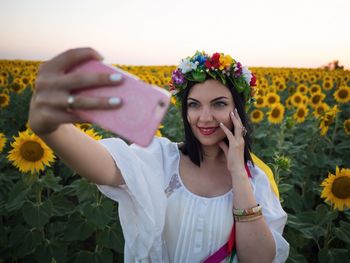 Smiling woman wearing flowers taking selfie through mobile phone while standing on field