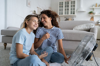 Overjoyed mother and teen daughter enjoying fresh cold air from electrical fan, sitting on floor