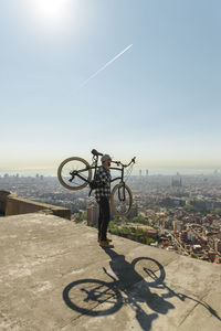 Side view of man carrying bicycle while standing on retaining wall over cityscape against sky during sunny day