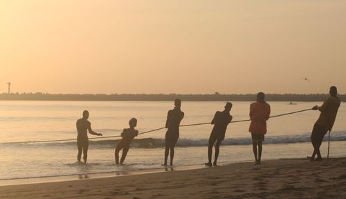 Silhouette people pulling rope on shore at beach against sky during sunset