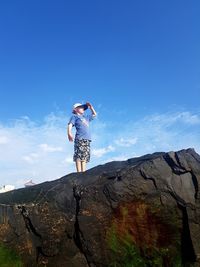 Low angle view of boy standing on rocky mountain against blue sky