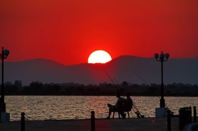 Silhouette people sitting on shore against orange sunset sky