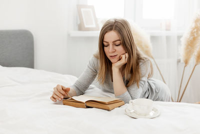 Young girl reading a book and daydreaming while sitting on the bed at home