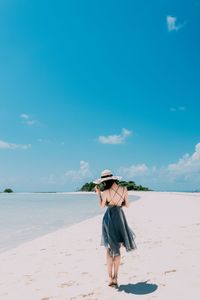 Rear view of young woman standing at beach against blue sky during sunny day