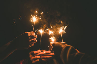 Cropped hands of people holding sparklers against sky at night