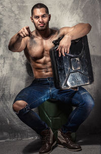 Full length portrait of shirtless muscular man with gas can pointing against wall