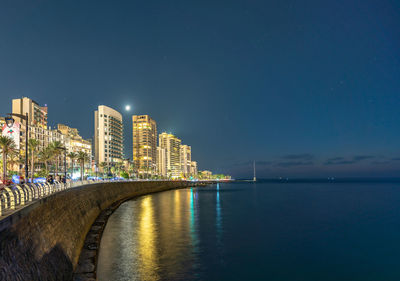 Illuminated buildings by sea against sky at night