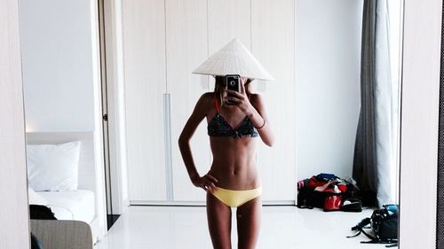 Reflection of woman in bikini with asian style conical hat taking selfie at home