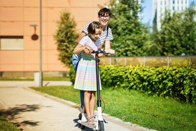 Mother with daughter riding push scooter on footpath