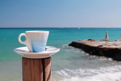 Close-up of coffee cup on wooden post at beach against sky