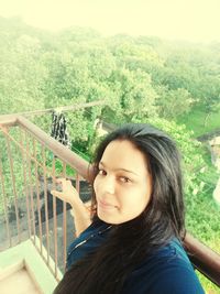 Portrait of beautiful woman standing by railing against trees