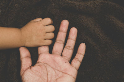 High angle view of baby holding finger of parent on brown blanket