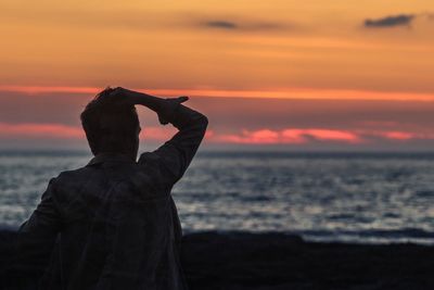 Rear view of man with hand in hair standing at beach during sunset