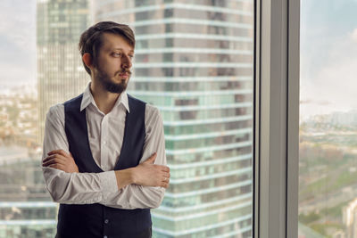 Thoughtful businessman with beard stands next to  window in office backdrop of skyscrapers in moscow