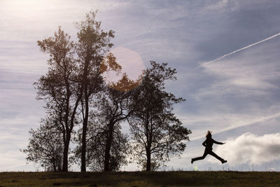 Silhouette woman jumping by trees on field against sky