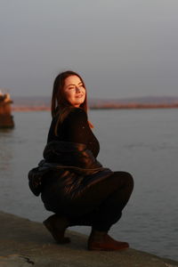 Woman crouching by sea against sky during sunset