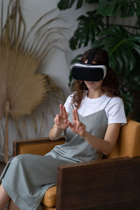 Curious woman in vr helmet touching ar interface with fingers, sitting in urban jungle interior