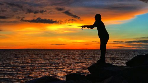 Silhouette man fishing in sea against cloudy sky during sunset