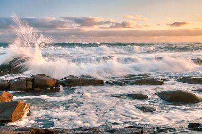 Waves splashing on rocks against sky during sunset