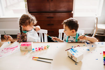 Brother and sister sitting at table painting with rainbow paints