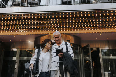 Smiling senior man sharing smart phone with female elderly friend outside movie theater