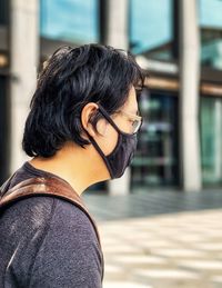 Side view of young asian man in eyeglasses and face mask against building exterior.