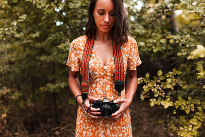 Young woman holding camera while standing against trees in forest