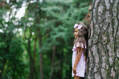 Portrait of a cute little girl leaning against a big tree with her back in the forest in summer