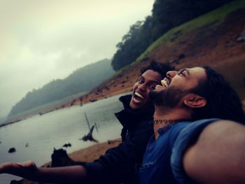 Tilt shot of cheerful men at lakeshore against sky