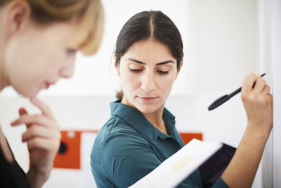 Mid adult businesswoman writing on whiteboard while colleague reading document in office