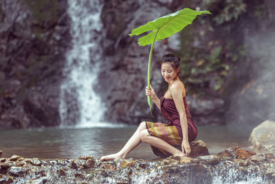 Portrait of young woman sitting on rock against waterfall