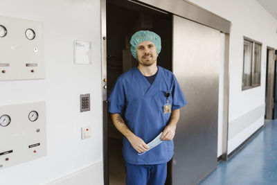 Portrait of smiling physician wearing scrubs standing at doorway in hospital corridor