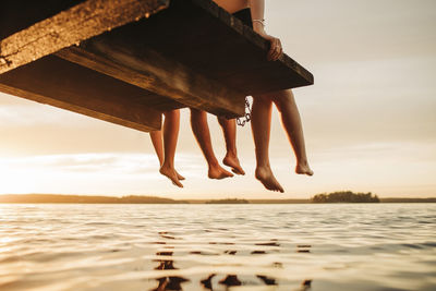 Low section of male and female friends sitting on jetty by lake during sunset