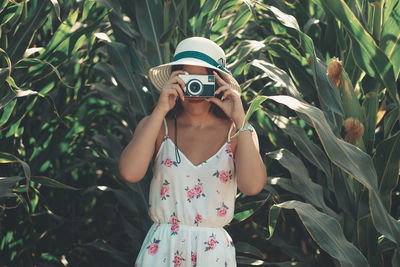 Young woman standing against plants taking a photo with a vintage camera