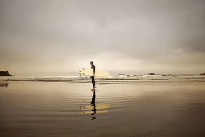 Side view of man carrying surfboard while standing on beach against sky