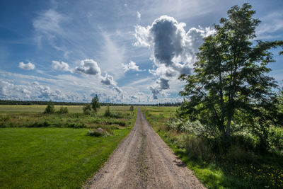 Dirt road amidst green field against cloudy sky during sunny day