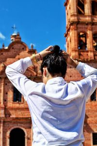 Rear view of young woman tying hair while standing by temple against sky